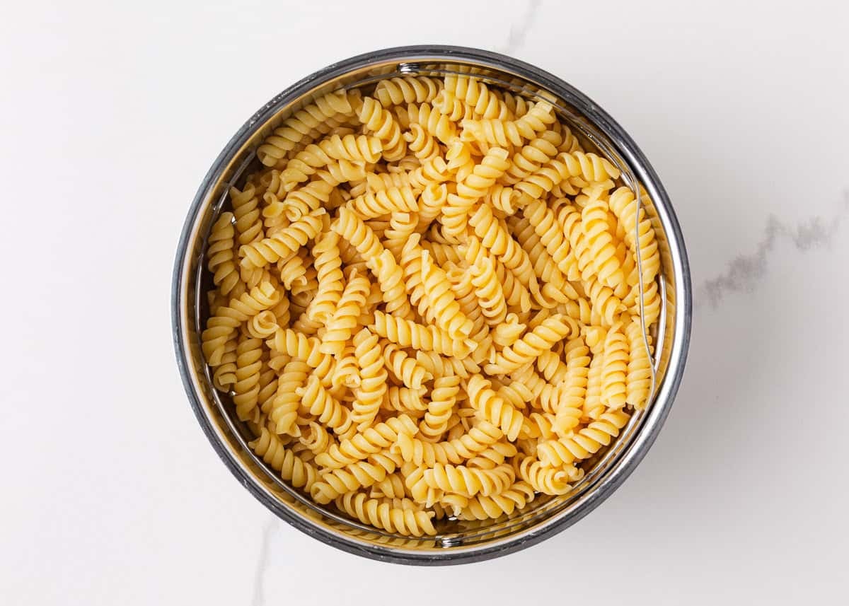 Packed with bold flavors in every bite, this cowboy pasta salad is made with tender noodles, bacon, seasoned ground beef, a creamy dressing, and Southwestern flavor! Cooked rotini pasta draining in a metal colander after rinsing.