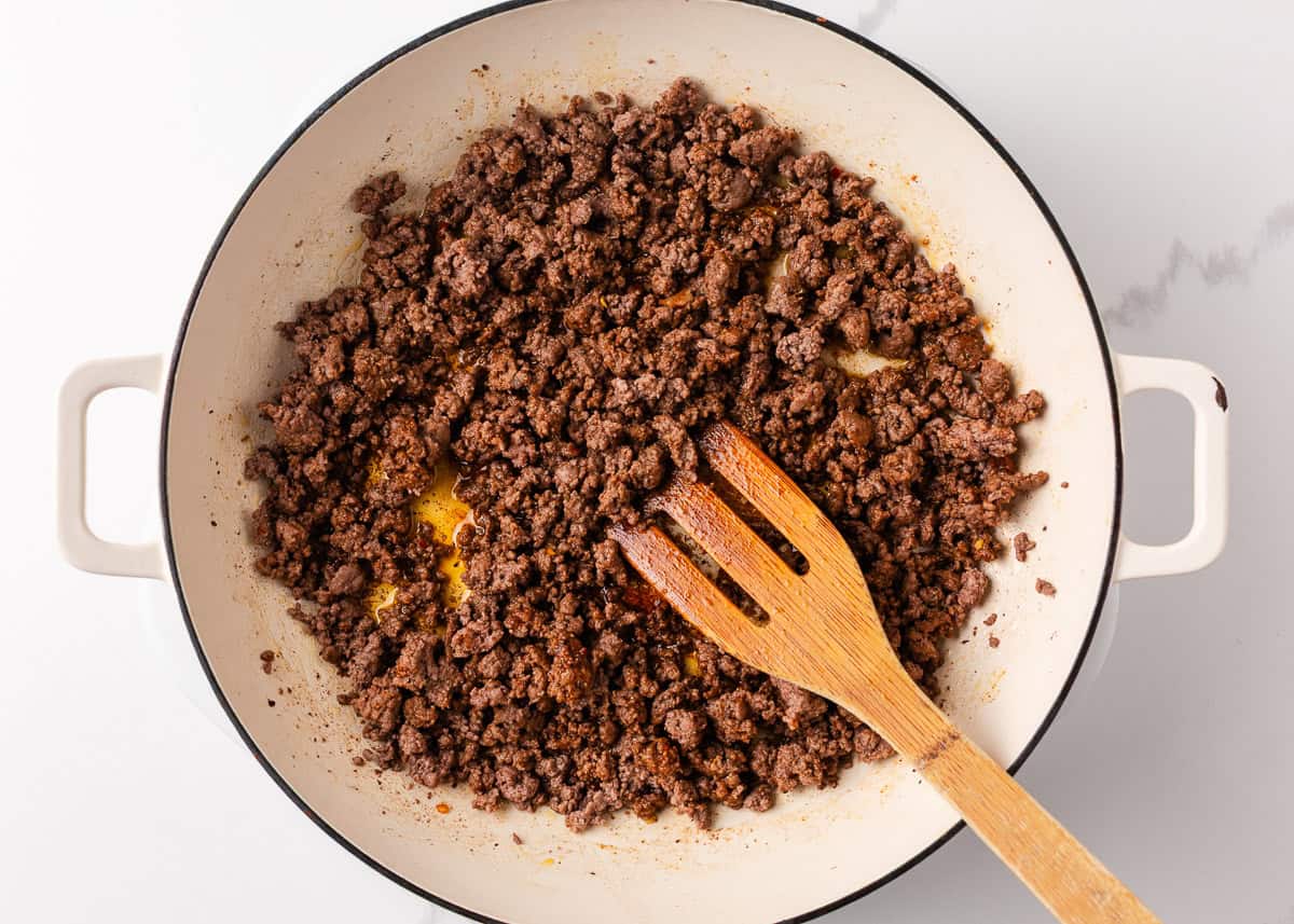 Packed with bold flavors in every bite, this cowboy pasta salad is made with tender noodles, bacon, seasoned ground beef, a creamy dressing, and Southwestern flavor! Seasoned ground beef browning in a Dutch oven, broken into crumbles with a wooden spatula.