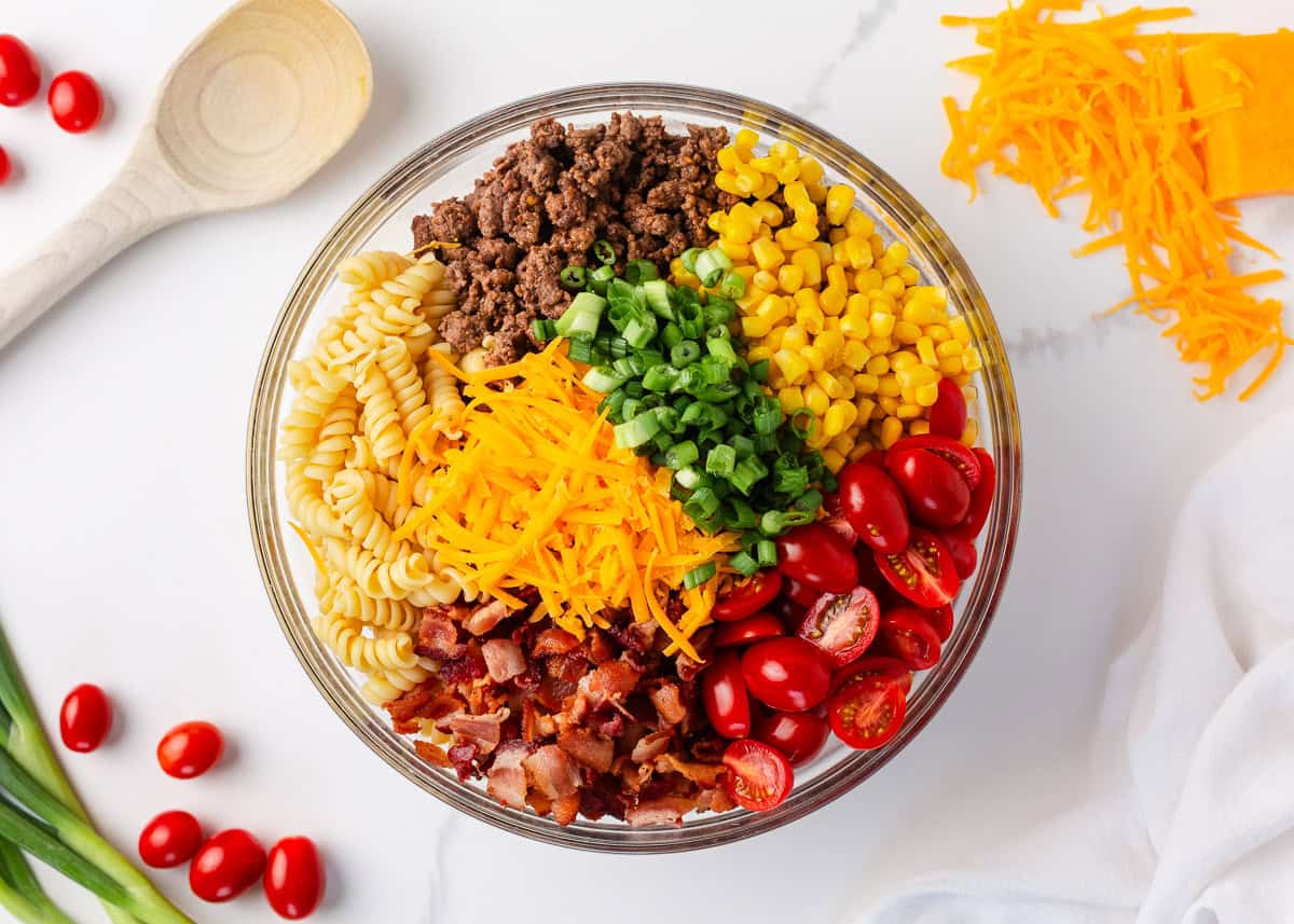 Packed with bold flavors in every bite, this cowboy pasta salad is made with tender noodles, bacon, seasoned ground beef, a creamy dressing, and Southwestern flavor! Cowboy pasta salad ingredients arranged in sections in a large glass bowl before tossing.
