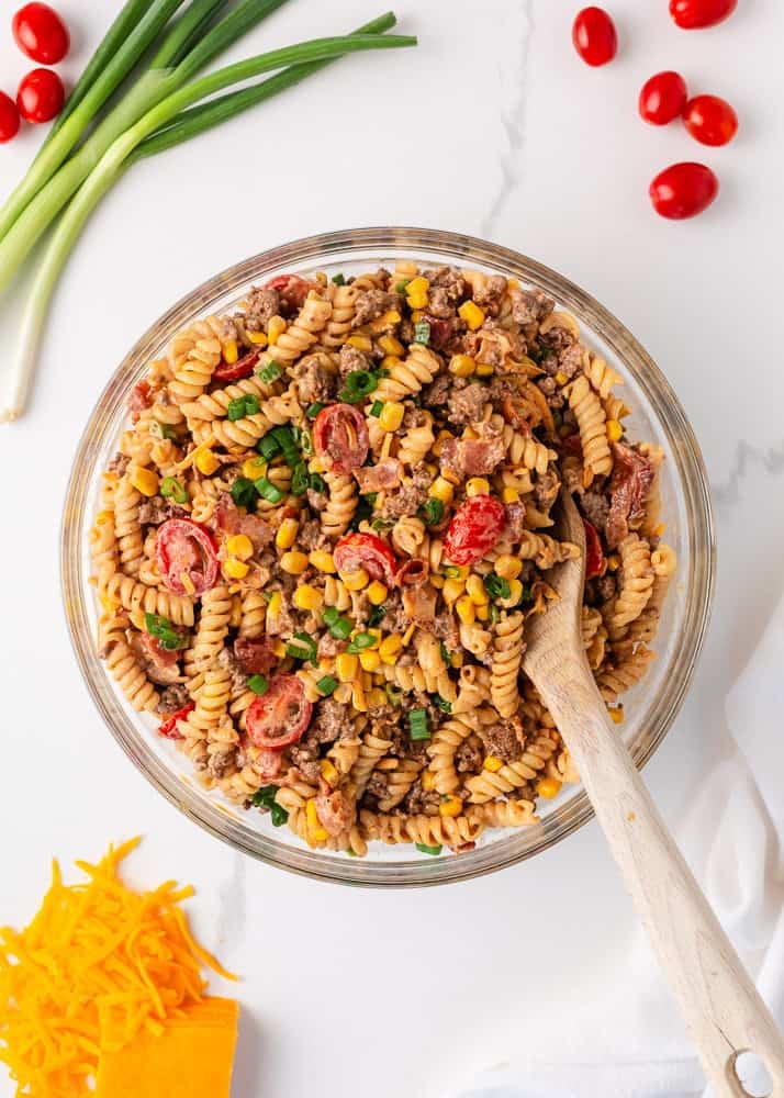 Packed with bold flavors in every bite, this cowboy pasta salad is made with tender noodles, bacon, seasoned ground beef, a creamy dressing, and Southwestern flavor! Overhead view of the finished cowboy pasta salad tossed in a large glass bowl with a wooden spoon.