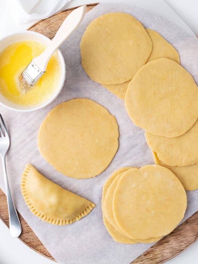 Rolled out empanada dough discs on parchment paper with egg wash and a pastry brush.