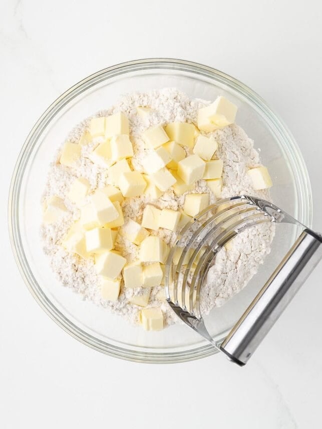 Cubed cold butter and flour in a glass bowl with a pastry cutter for making empanada dough.