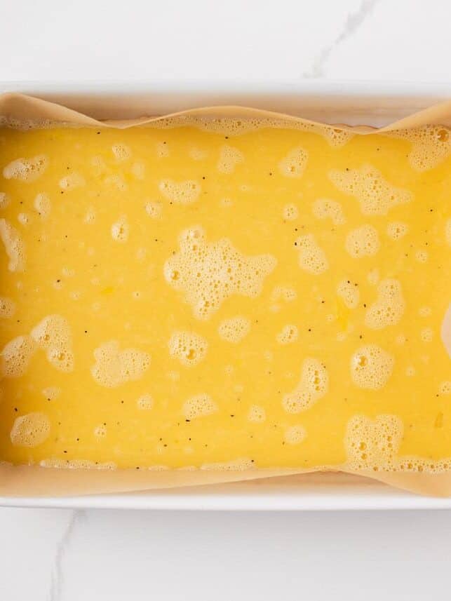 Overhead view of a white baking dish lined with parchment paper, filled with a seasoned egg mixture ready to be baked into a sheet egg for breakfast sliders.