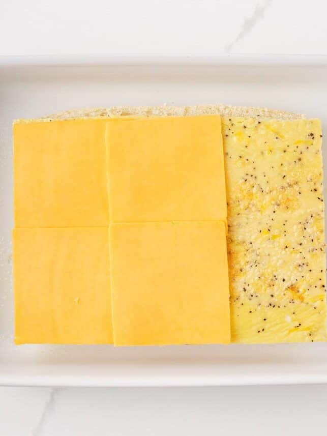 Overhead view of sliced cheddar cheese placed on top of a baked sheet egg on the bottom half of Hawaiian rolls in a white baking dish.