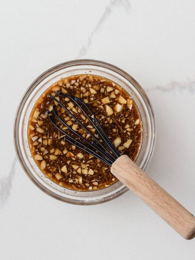 This pineapple chicken is sweet, tangy, and on the table in 30 minutes. Tender chicken, crisp bell peppers, and juicy pineapple in a sauce that beats takeout every time. Overhead view of pineapple chicken sauce with soy sauce, honey, garlic, and ginger whisked together in a glass bowl with a small black whisk.