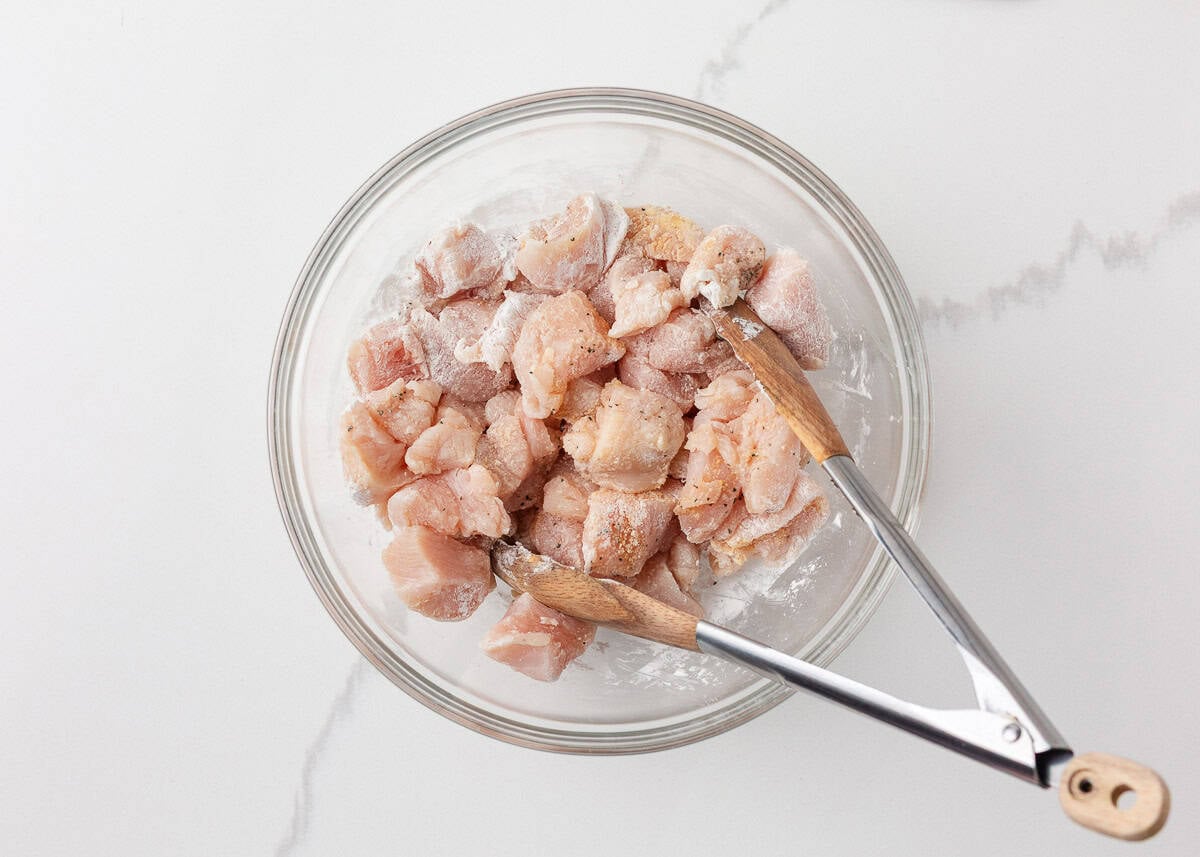 This pineapple chicken is sweet, tangy, and on the table in 30 minutes. Tender chicken, crisp bell peppers, and juicy pineapple in a sauce that beats takeout every time. Overhead view of raw cubed chicken breast tossed with cornstarch, salt, and pepper in a glass mixing bowl with tongs.