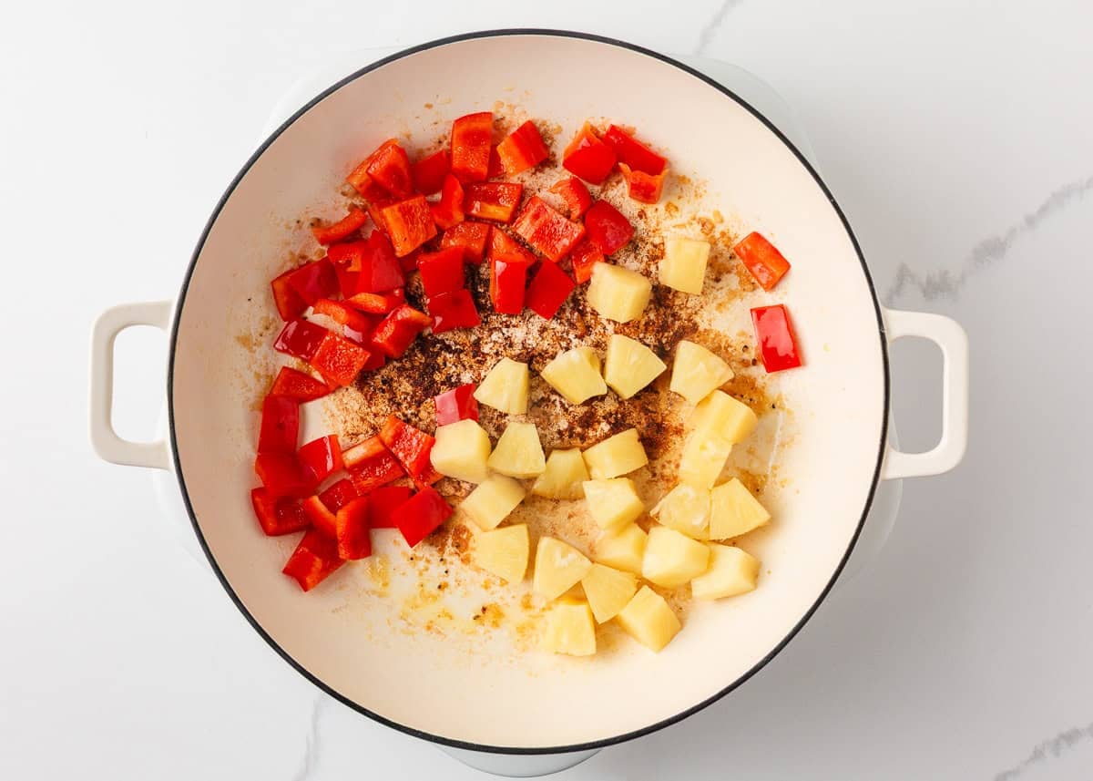 This pineapple chicken is sweet, tangy, and on the table in 30 minutes. Tender chicken, crisp bell peppers, and juicy pineapple in a sauce that beats takeout every time. Overhead view of chopped red bell pepper and pineapple chunks added to a white skillet during the cooking process.