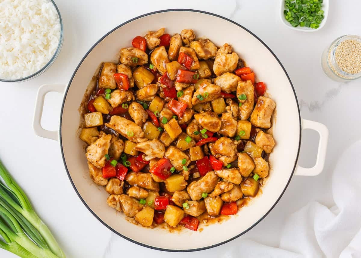 This pineapple chicken is sweet, tangy, and on the table in 30 minutes. Tender chicken, crisp bell peppers, and juicy pineapple in a sauce that beats takeout every time. Overhead view of completed pineapple chicken in a white skillet, with chicken, pineapple chunks, and red bell pepper coated in a glossy sauce and garnished with sliced green onions, surrounded by bowls of rice, sesame seeds, and green onions.