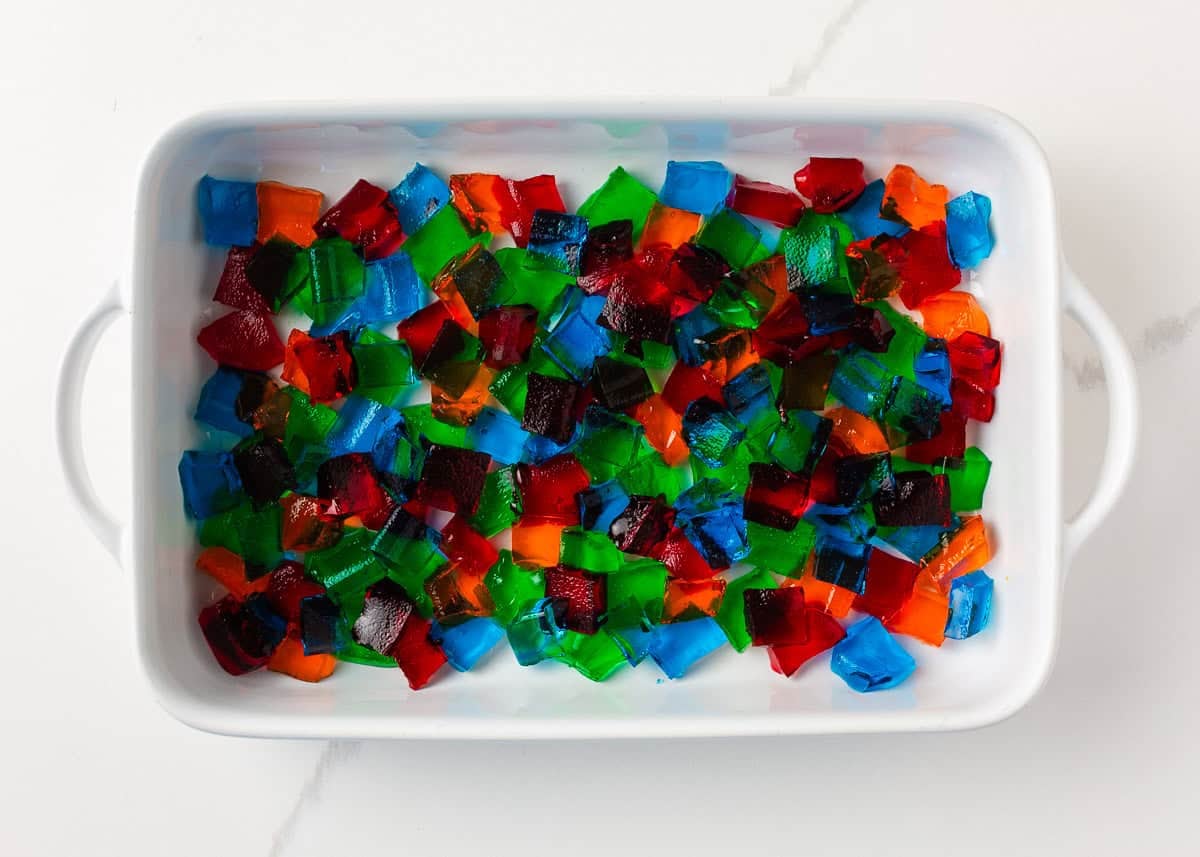 Made with Jello and sweetened condensed milk, stained glass jello has a fun, brightly colored, broken glass look and is the perfect make-ahead dessert for all your parties! Colorful cubes of red, blue, green, and orange Jell-O scattered in a white baking dish, showing how to make broken glass jello before adding the condensed milk mixture.