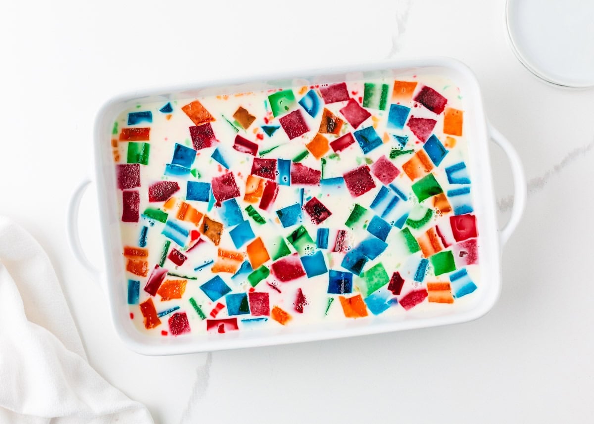 Made with Jello and sweetened condensed milk, stained glass jello has a fun, brightly colored, broken glass look and is the perfect make-ahead dessert for all your parties! The condensed milk gelatin poured over multicolored Jell-O cubes in a white handled baking dish, showing the final assembly step for stained glass jello salad before chilling.