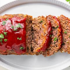 Sliced meatloaf on a white platter with glossy red meatloaf sauce and fresh parsley.