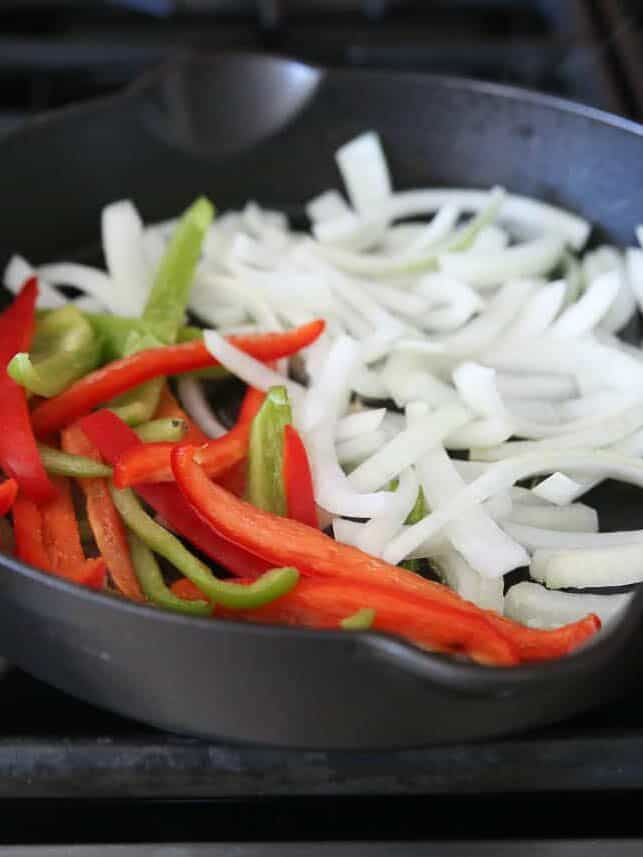 Sliced raw red and green bell peppers and white onion in a black cast iron skillet before cooking.