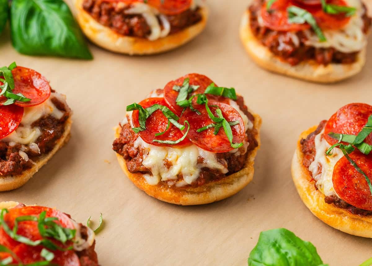 Bite into simpler times with old-school open faced pizza burgers made with seasoned ground beef on a toasted bun, smothered in tomato sauce, melty cheese, and pepperoni! Close-up of open-faced pizza burgers with melted cheese, pepperoni slices, and fresh basil on toasted buns.