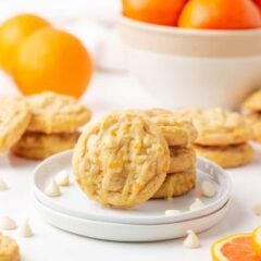 Orange creamsicle cookies on a plate with fresh oranges in the background and white chocolate chips scattered around.