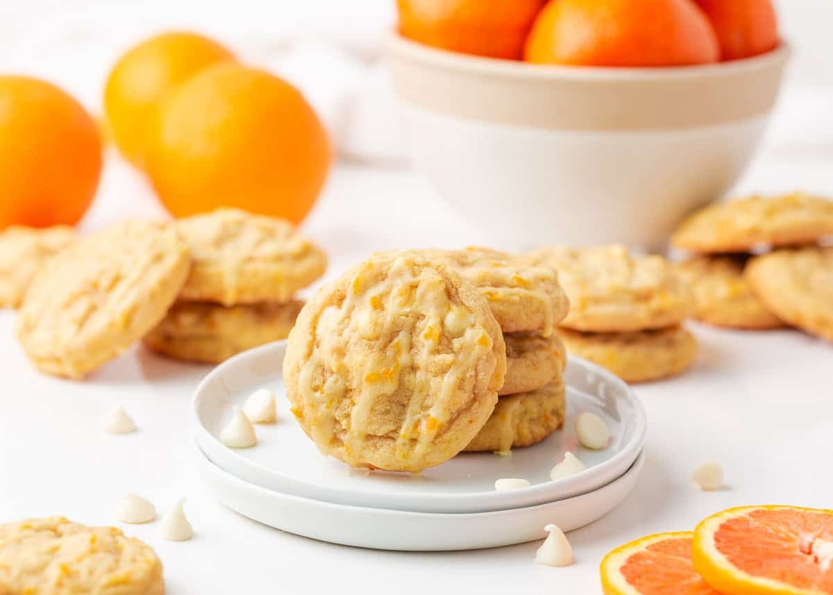 Orange creamsicle cookies on a plate with fresh oranges in the background and white chocolate chips scattered around.