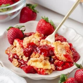 Serving of strawberry cheesecake dump cake on plate with fresh strawberries and crumb cake topping.