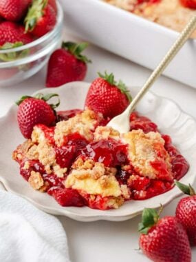 Serving of strawberry cheesecake dump cake on plate with fresh strawberries and crumb cake topping.