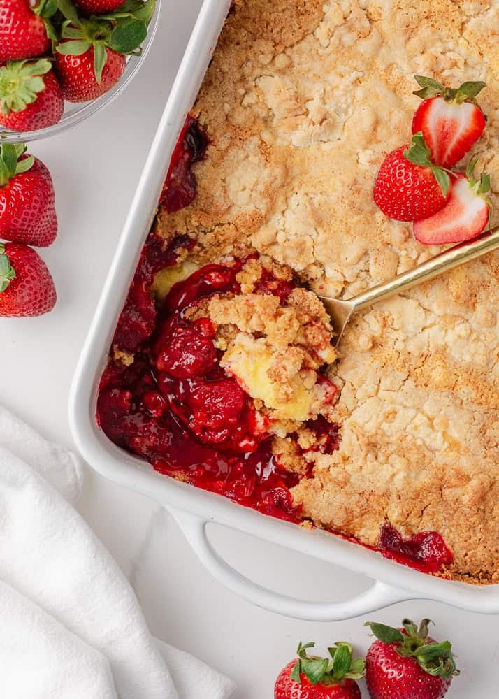 Easy and sweet strawberry cheesecake dump cake is made with strawberry pie filling, cream cheese, boxed cake mix, and just 5 minutes of prep time! Spoon scooping strawberry cheesecake dump cake with golden crumb topping and gooey strawberry filling from white baking dish.