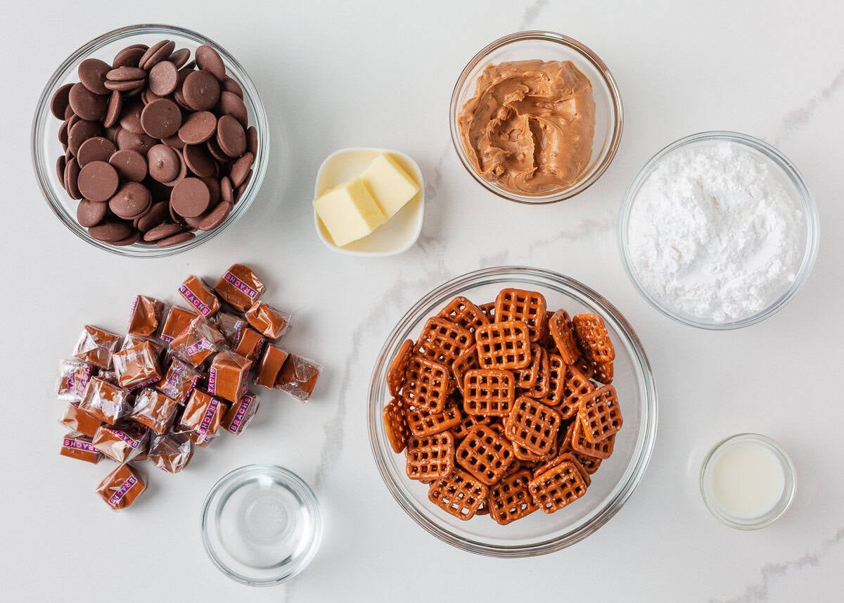 If you love Take 5 candy bars, you are going to love this easy homemade version. No baking required and every bite is the perfect mix of salty, sweet, and crunchy. Overhead flat lay of ingredients in glass bowls on a white marble surface: chocolate melting wafers, crunchy peanut butter, softened butter, powdered sugar, wrapped caramel squares, square pretzel snaps, water, and milk.