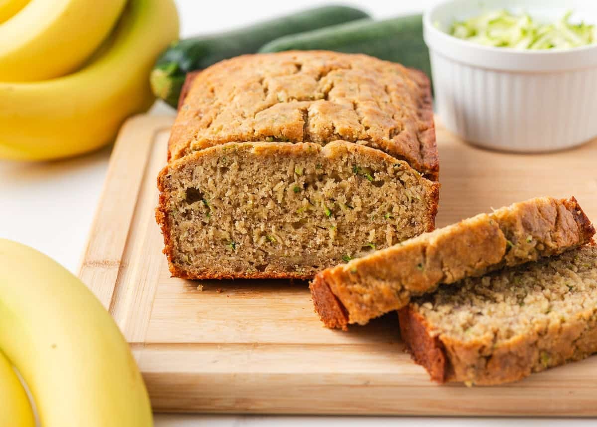Moist, tender, and packed with flavor, this zucchini banana bread is a delicious way to use up ripe bananas and fresh zucchini in one easy loaf! Sliced zucchini banana bread loaf on a cutting board with zucchini and bananas in the background.