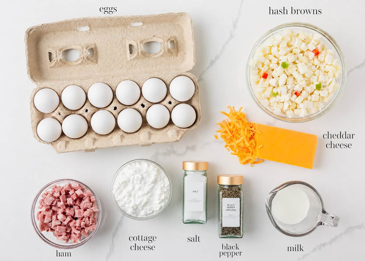 Ingredients for Amish breakfast casserole arranged on a white marble surface, including a carton of 12 eggs, a bowl of frozen potatoes O'Brien, shredded and block cheddar cheese, diced ham, small curd cottage cheese, salt, black pepper, and a glass measuring cup of milk.