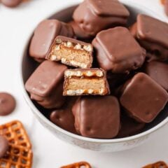 Overhead view of a white bowl filled with chocolate-dipped pretzel bites, two of which are cut in half showing the peanut butter and caramel layers, surrounded by loose pretzel snaps and chocolate wafers.