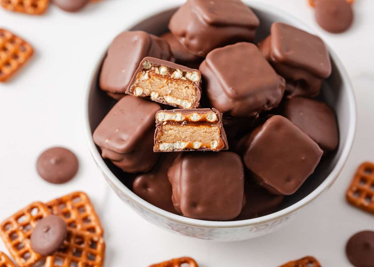If you love Take 5 candy bars, you are going to love this easy homemade version. No baking required and every bite is the perfect mix of salty, sweet, and crunchy. Overhead view of a white bowl filled with chocolate-dipped pretzel bites, two of which are cut in half showing the peanut butter and caramel layers, surrounded by loose pretzel snaps and chocolate wafers.