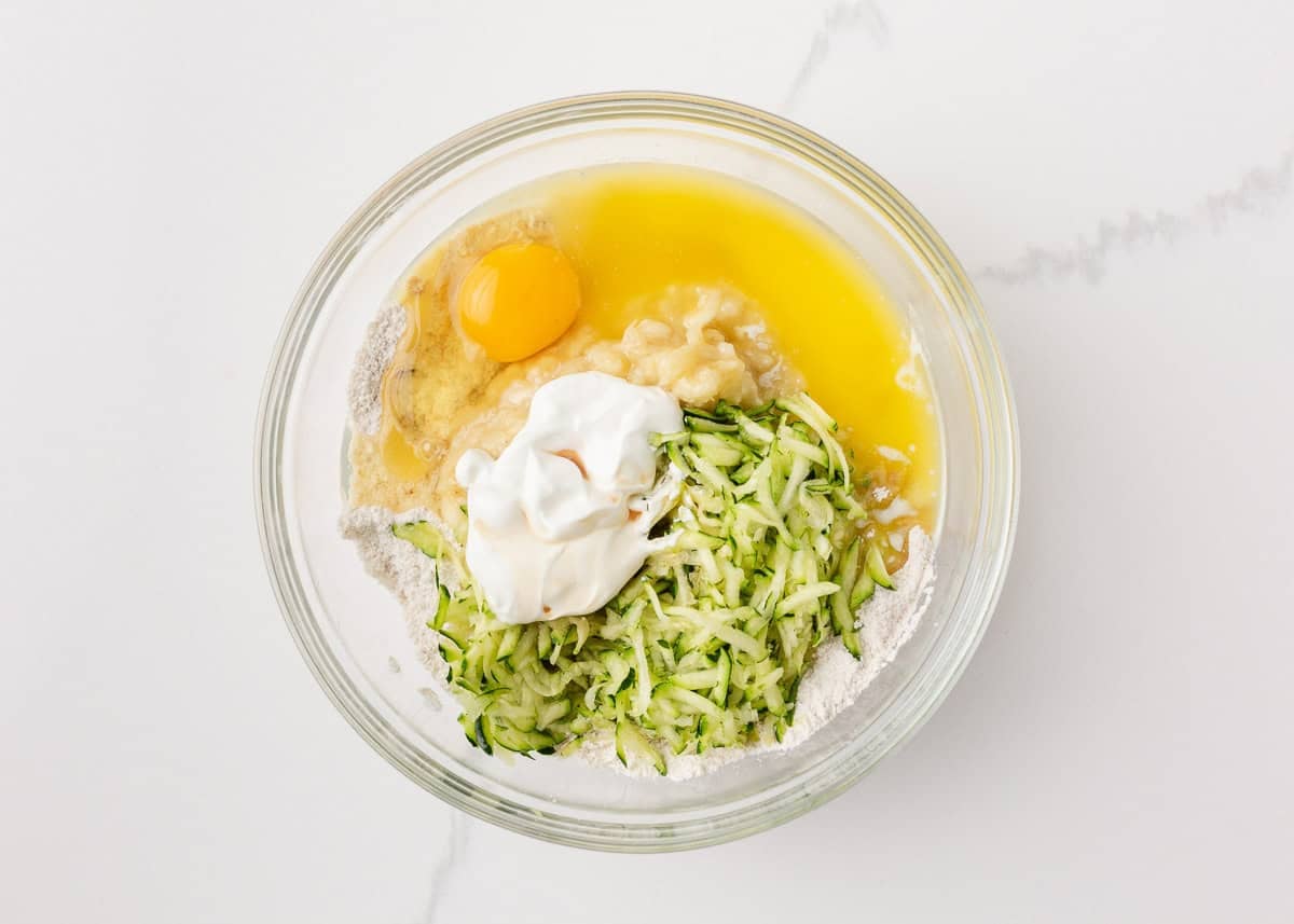 Moist, tender, and packed with flavor, this zucchini banana bread is a delicious way to use up ripe bananas and fresh zucchini in one easy loaf! Zucchini banana bread ingredients including grated zucchini, mashed banana, egg, sour cream and melted butter in a glass bowl.