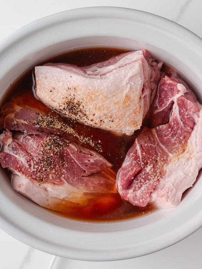 Raw pork shoulder in a slow cooker with barbecue sauce and seasonings before cooking.