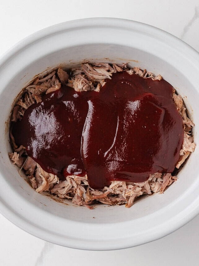 Shredded pork topped with barbecue sauce in a slow cooker for BBQ pulled pork sandwiches.