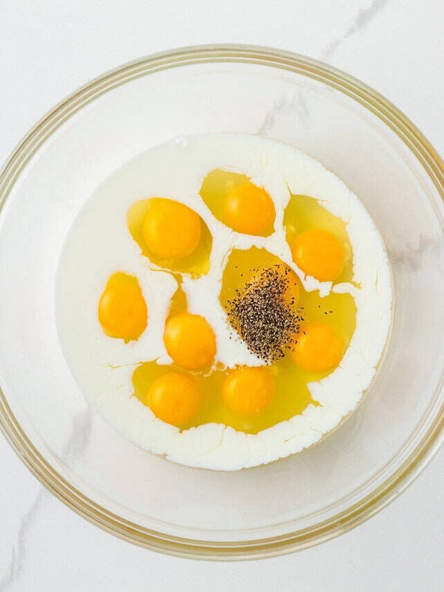 Overhead view of nine cracked eggs with milk and black pepper in a large glass mixing bowl, before whisking.