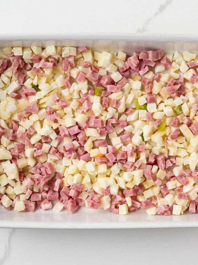 Overhead view of a white 9x13-inch baking dish evenly layered with frozen potatoes O'Brien and diced ham, ready for the egg mixture to be poured over.