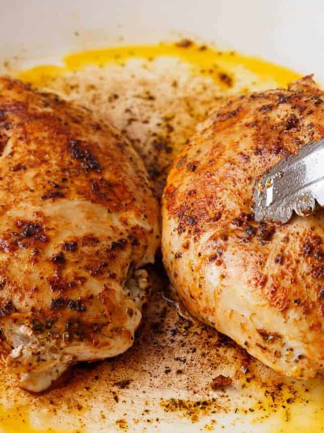Two seasoned chicken breasts cooking in a skillet with tongs.