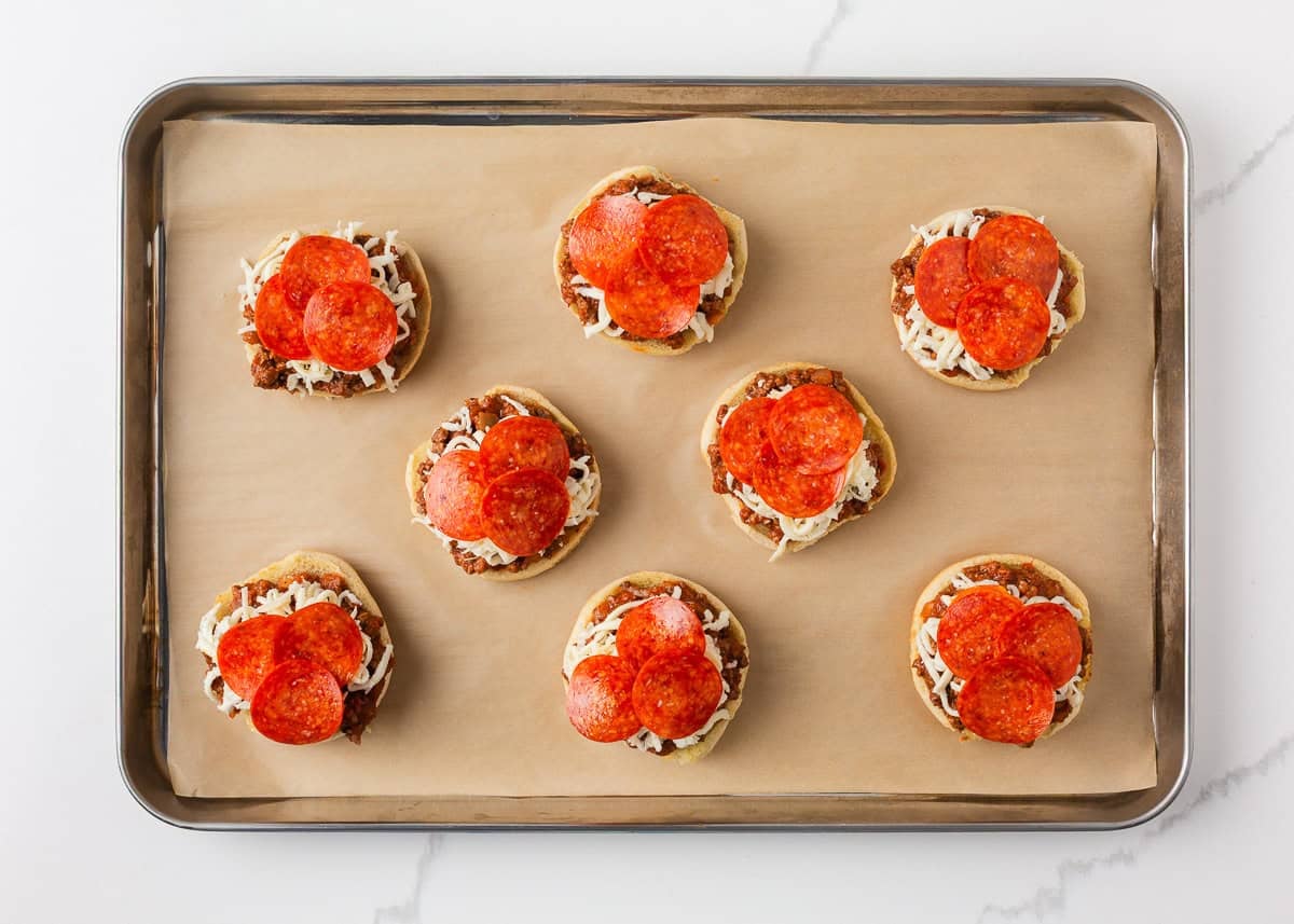 Bite into simpler times with old-school open faced pizza burgers made with seasoned ground beef on a toasted bun, smothered in tomato sauce, melty cheese, and pepperoni! Open-faced pizza burgers assembled on a lined baking sheet before baking with cheese and pepperoni topping.