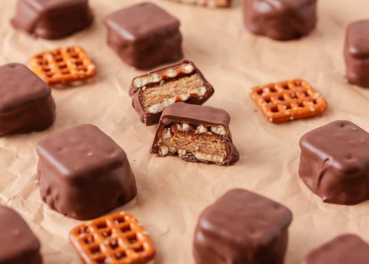 If you love Take 5 candy bars, you are going to love this easy homemade version. No baking required and every bite is the perfect mix of salty, sweet, and crunchy. Several finished chocolate-dipped bites scattered on brown parchment paper alongside loose pretzel snaps, one bite cut open to show the layered interior.