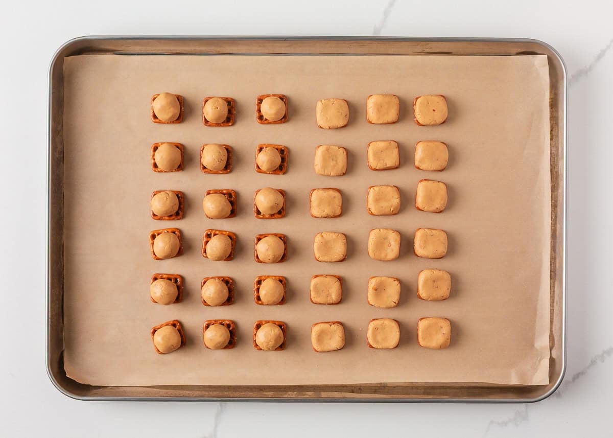 If you love Take 5 candy bars, you are going to love this easy homemade version. No baking required and every bite is the perfect mix of salty, sweet, and crunchy. Overhead shot of a baking sheet lined with parchment showing two sections: one with peanut butter balls pressed onto pretzel snaps, and one with the flattened peanut butter-pretzel sandwiches ready to be topped.