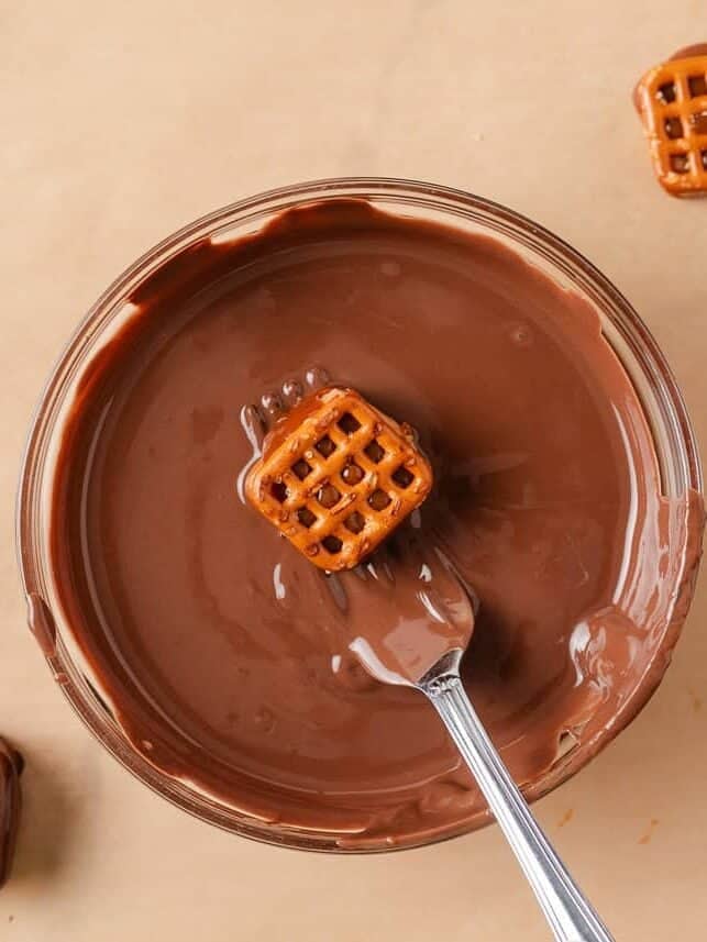 If you love Take 5 candy bars, you are going to love this easy homemade version. No baking required and every bite is the perfect mix of salty, sweet, and crunchy. Overhead shot of a pretzel bite being lifted out of a bowl of melted chocolate on a fork, with partially dipped bites visible on parchment paper around the bowl.