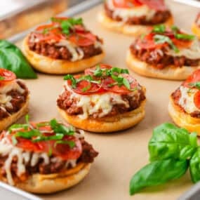 Pizza burgers on a baking sheet topped with beefy pizza mixture, melted cheese, pepperoni slices, and fresh basil.