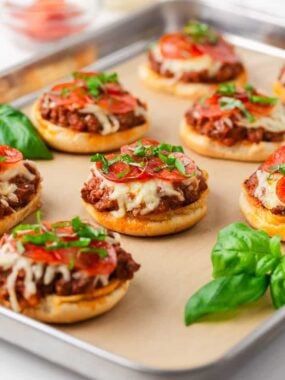 Pizza burgers on a baking sheet topped with beefy pizza mixture, melted cheese, pepperoni slices, and fresh basil.