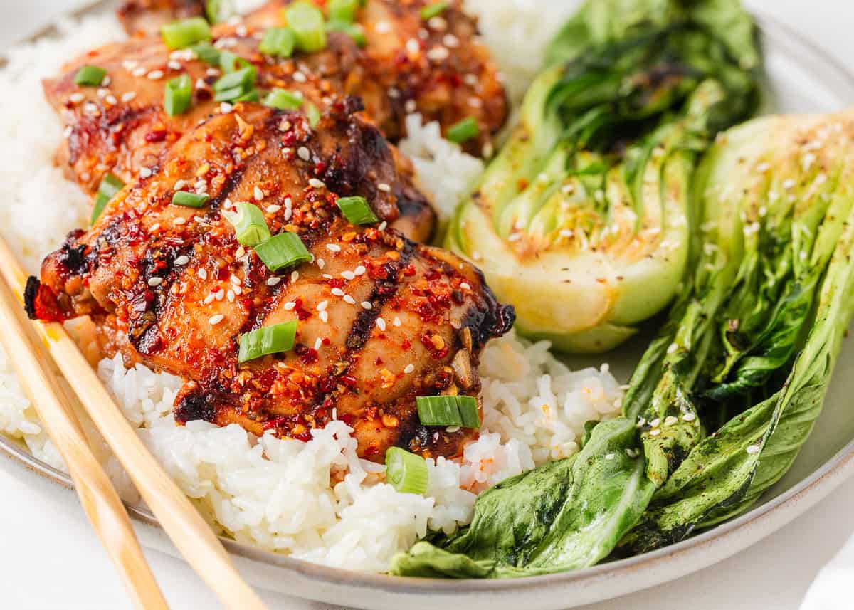 Close up of Asian chicken thighs with chili crunch, sesame seeds and grilled bok choy over white rice.