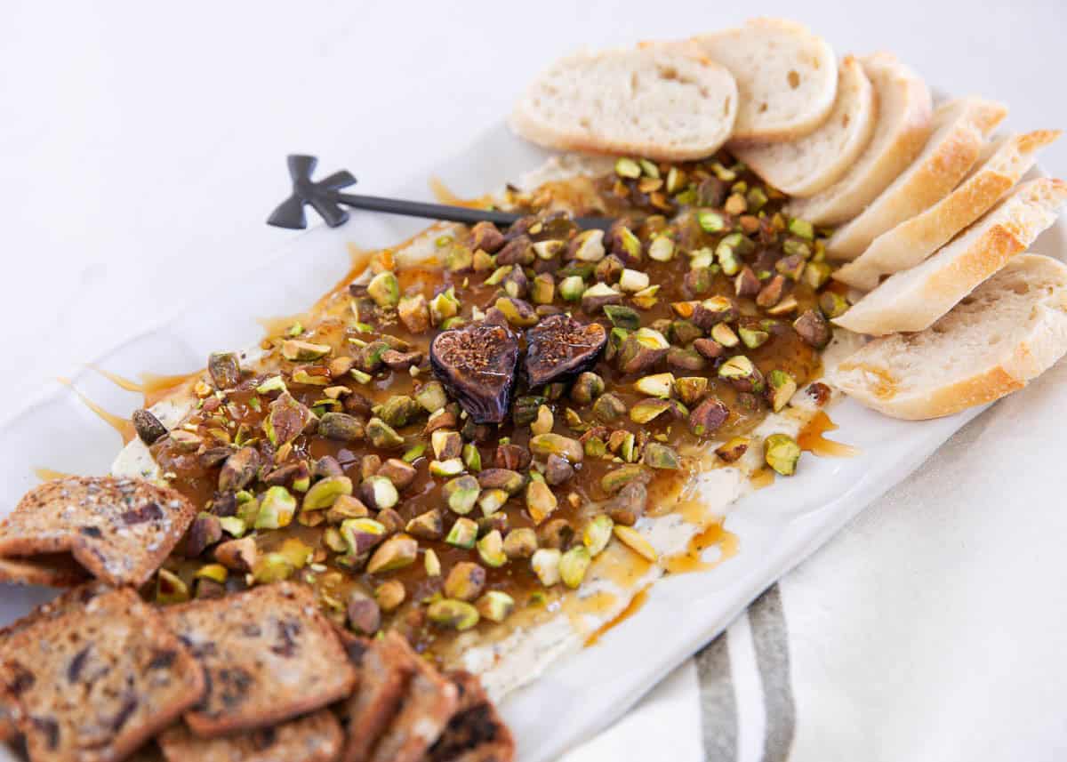 Boursin fig dip with pistachios and hot honey served on a platter with sliced baguette and crackers for dipping.