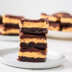 Close up of three buckeye brownies stacked on a white plate with a rich peanut butter layer and chocolate topping.