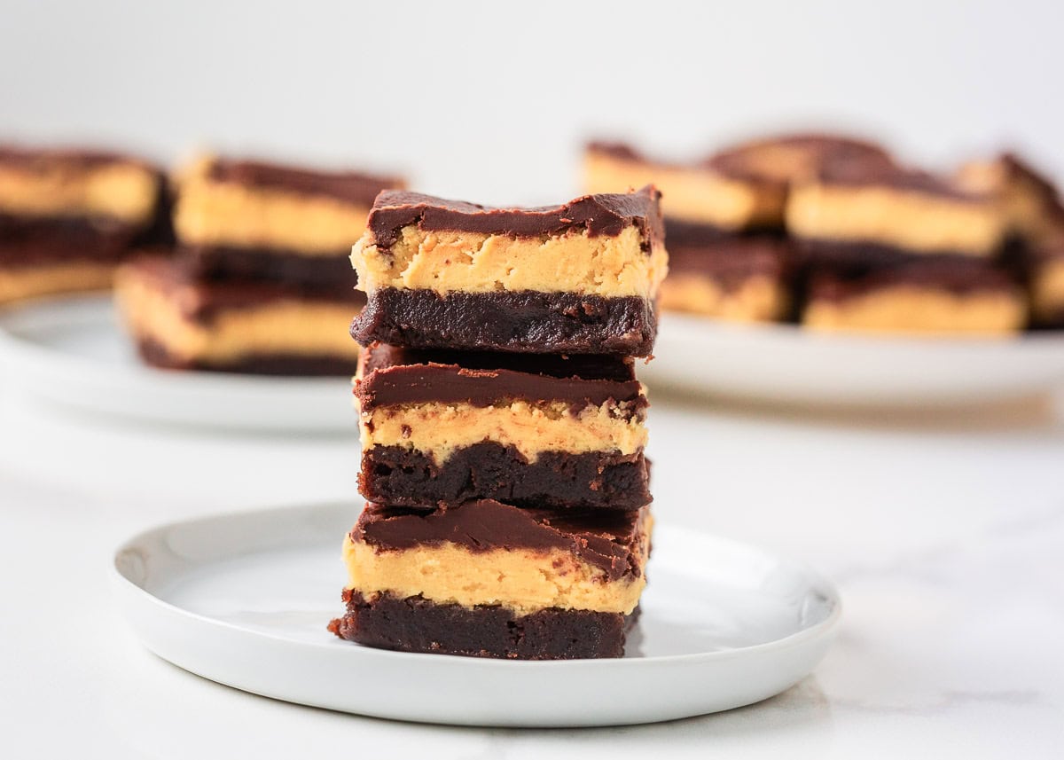 Close up of three buckeye brownies stacked on a white plate with a rich peanut butter layer and chocolate topping.