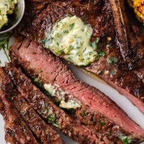 Sliced grilled steak with garlic herb butter melting over the top and grilled corn on the side.