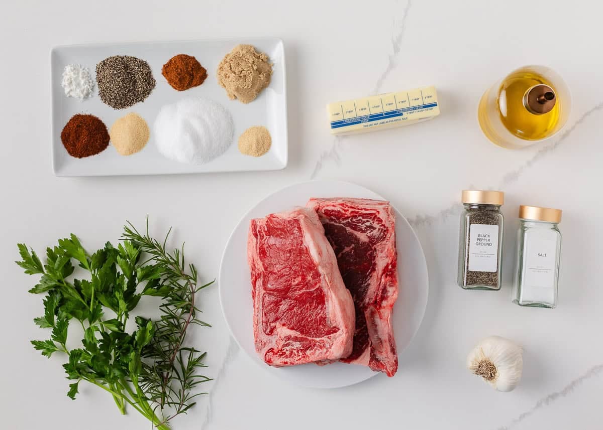 Grilled steak ingredients with bone-in steaks, homemade steak rub, garlic, herbs, butter and oil on a marble counter.