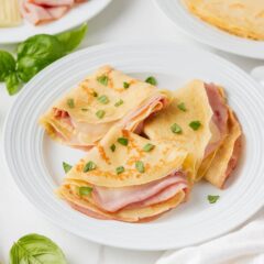 Three folded ham and cheese crepes stacked on a white plate with fresh basil.