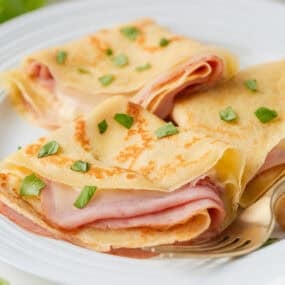 Ham and cheese crepes folded on a white plate with fresh basil and gold fork.