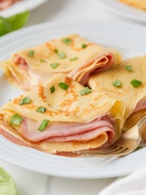 Ham and cheese crepes folded on a white plate with fresh basil and gold fork.