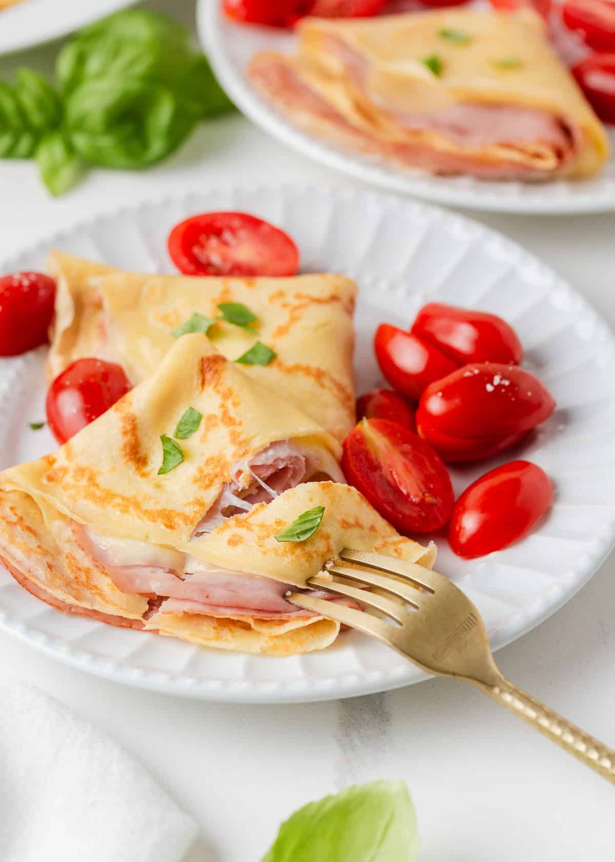 Ham and cheese crepes are made with slices of delicious ham and melted cheese and served with the option of juicy cherry tomatoes. A delicious savory crepe that's so simply made, it's perfect for breakfast, lunch, dinner, and everything in between! Ham and cheese crepe on a white plate with fresh cherry tomatoes and basil.