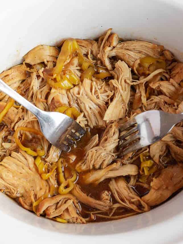 Two forks shredding tender Mississippi chicken in the crockpot with pepperoncini peppers and buttery au jus sauce.